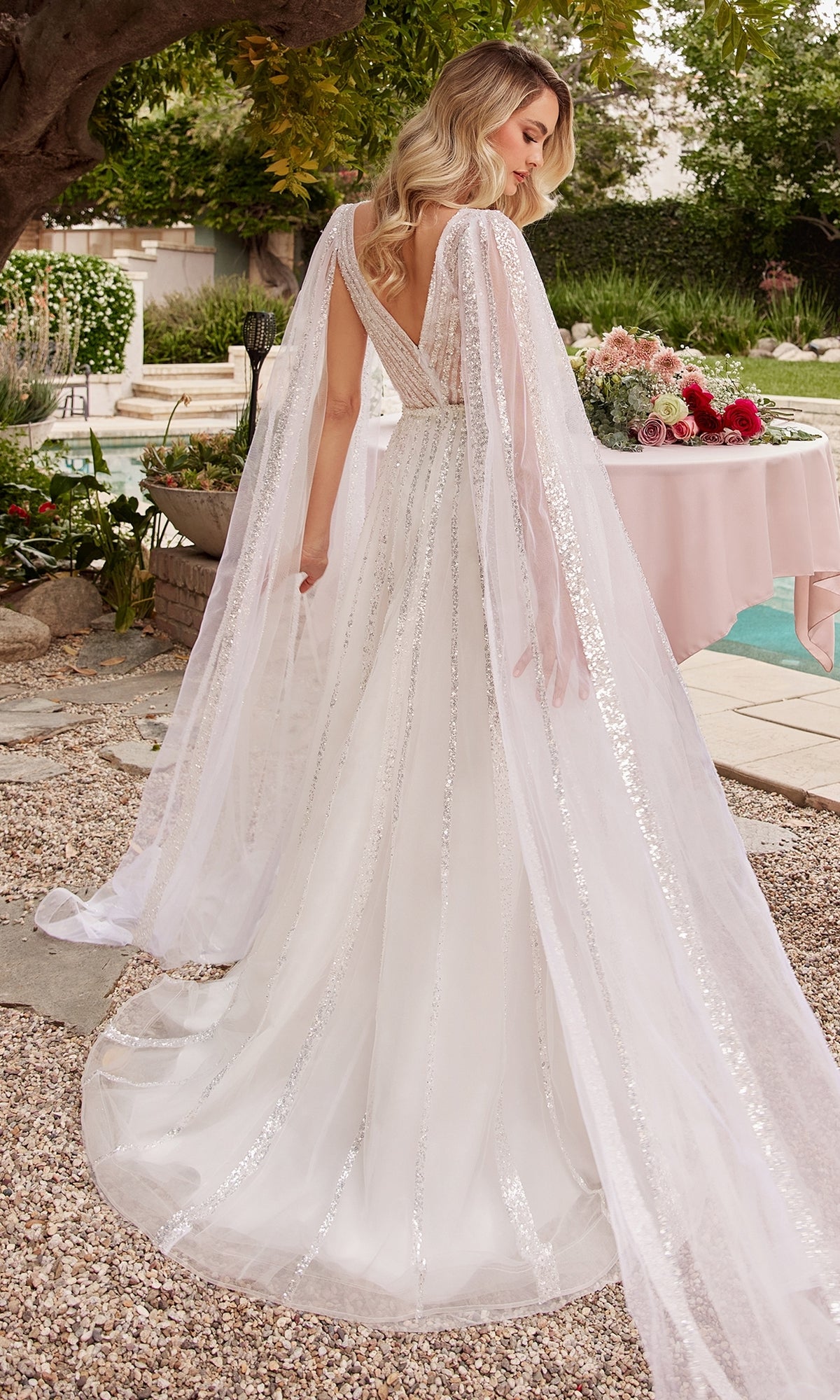A woman wears the Ladivine CD852W Drape-Sleeve White Formal Dress with sequins, deep V-back, and sheer cape, standing outdoors by a flower-adorned table.